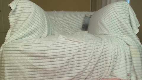 Media: A video of a white, textured, ribbed blanket draped over a chair, creating a cozy, warm atmosphere. The background is a beige wall with a partially visible window and white curtains.