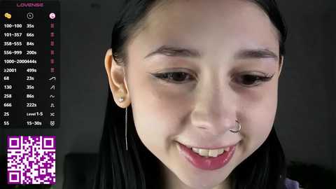 Video of a young Asian woman with long black hair, wearing a nose ring, smiling. Background includes a camera viewfinder and a QR code.