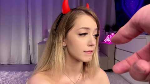 Video of a young blonde woman with light skin, wearing red devil horns, a white top, and a necklace, sitting in a dimly lit room with a man's hand caressing her cheek.