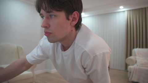 Video of a young, fair-skinned, dark-haired man in a white t-shirt, leaning forward in a modern living room with beige furniture, white walls, and soft lighting.