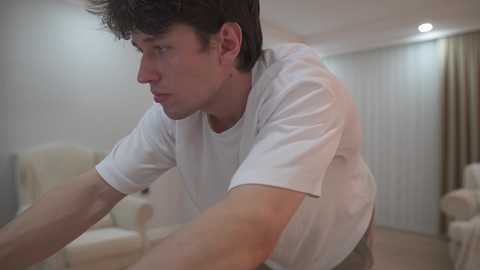Video of a young man with short dark hair in a white t-shirt, leaning forward intensely, in a dimly lit room with beige furniture and white curtains.