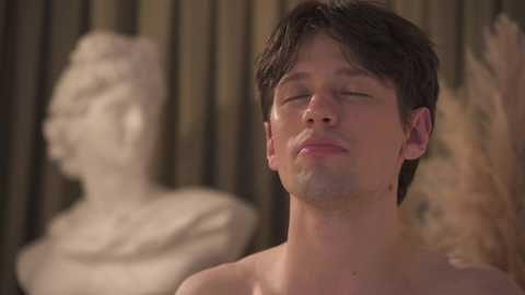 Video of a shirtless young man with short dark hair, closed eyes, and a relaxed expression, standing in front of a blurred white bust and wooden panel backdrop.