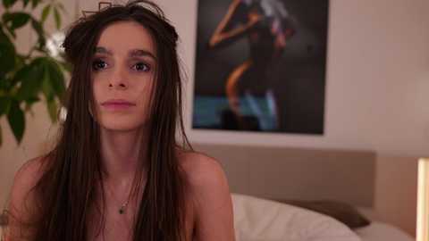 Video of a young woman with long, dark hair, wearing a delicate necklace, standing topless in a softly lit bedroom with a modern, abstract artwork in the background.