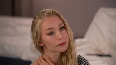 Video of a fair-skinned, blonde-haired young woman with light makeup, wearing a black-and-white checkered shirt, sitting on a bed with white sheets, looking directly at the camera with a neutral expression.