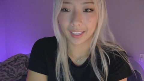 A video of an Asian woman with long, straight platinum blonde hair and fair skin, wearing a black t-shirt, smiling slightly. Background shows a blurred patterned fabric and purple lighting.