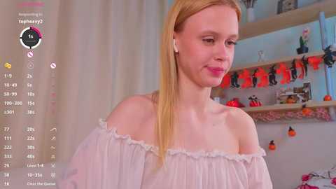 Video of a slender, fair-skinned woman with straight blonde hair, wearing a white off-shoulder blouse. Background features a shelf with decorative items, including a pumpkin, and a camera display.