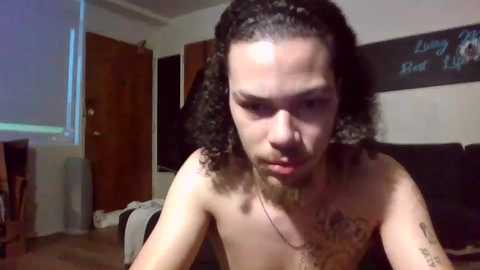 Video of a shirtless, light-skinned person with curly hair and tattoos, leaning forward, in a dimly lit room with a wooden door and black wall.