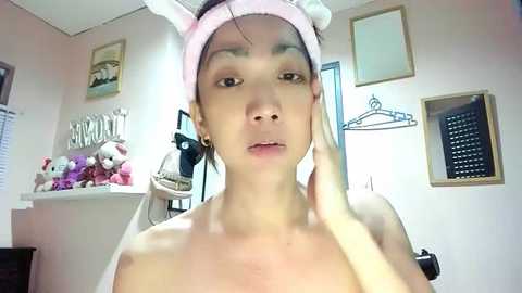 Media: Video of a topless Asian woman with light skin and dark hair, wearing a pink bunny headband, touching her face in a bathroom with pink walls and stuffed animals.
