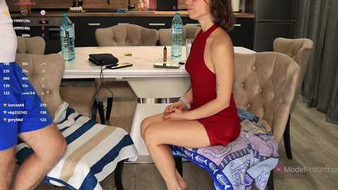 Video of a woman in a red dress sitting on a striped towel, next to a man in blue shorts, in a modern kitchen with beige chairs and water bottles.
