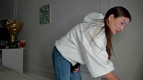 Video of a young woman with long brown hair, wearing a white hoodie and blue jeans, bending over in a modern living room.