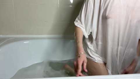 Video of a partially submerged, fair-skinned woman in a white t-shirt, sitting in a bathtub filled with water, in a bathroom with beige tiled walls.