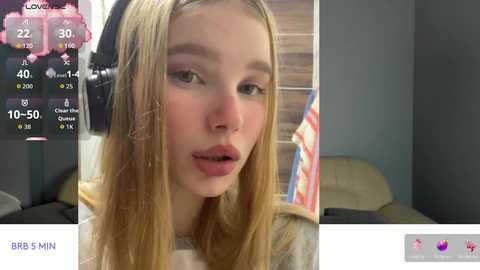 Video of a young Caucasian woman with fair skin and long blonde hair, wearing a grey sweater, captured indoors with a brick wall background.