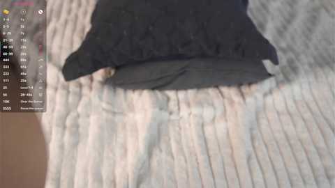 Media: A video showing a close-up of a person\u2019s hand and arm in a black sweater, resting on a light grey, textured sweater with ribbed edges, on a beige surface. The image includes a digital overlay with a grid and numerical data.