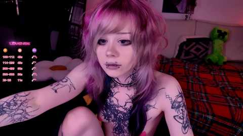 Video of a pale-skinned, androgynous individual with purple hair, wearing black lace lingerie, covered in tattoos, sitting on a bed with a plaid blanket, in a dimly lit room.
