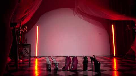 Media: Video of a dimly lit, abstract room with red and pink lighting, featuring a black-and-white checkered floor, two red candles, and a pile of high-heeled shoes.