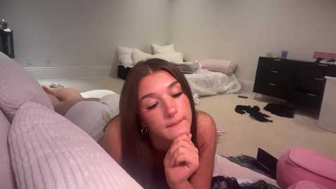 Video of a young, light-skinned woman with long brown hair, lying on her stomach in a messy bedroom, wearing a white top, performing oral sex on a man.