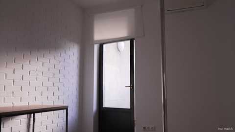 Video of a minimalist hallway with white brick walls, a black door with frosted glass, a wooden cabinet, and a metal chair, casting soft shadows.
