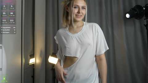 Video of a young woman with fair skin and blonde hair in a ponytail, wearing a sheer white T-shirt that reveals her breasts and nipples. She stands in a dimly lit room with grey curtains and a studio light in the background.