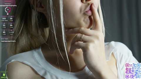 Video of a light-skinned woman with blonde hair, wearing a white off-the-shoulder top, touching her chin thoughtfully. Background shows a virtual reality headset with user stats.