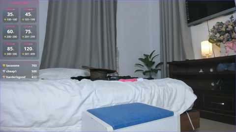 Video of a dimly lit bedroom with a white bed, blue cushion, grey curtains, dark wooden dresser, and digital weather display.