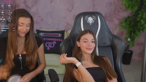 Video of two young women in a gaming room. One is braiding the other's hair; both wear casual black outfits. Background features gaming chairs, a plant, and gaming-themed decor.
