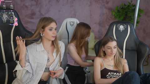Media: Video of three young women in a gaming room with black gaming chairs and a pink wall. Two women, one blonde, one brunette, are seated; the third, a brunette with a black crop top, is standing.
