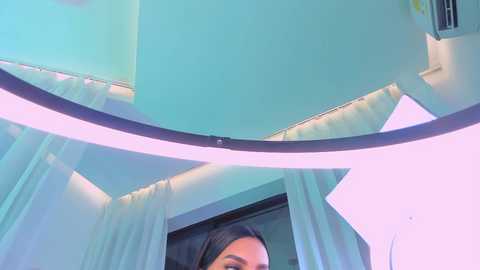 A video of a woman's face partially reflected in a curved, lit mirror, revealing a teal wall and soft pink lighting. The image is modern, with a focus on minimalist decor.