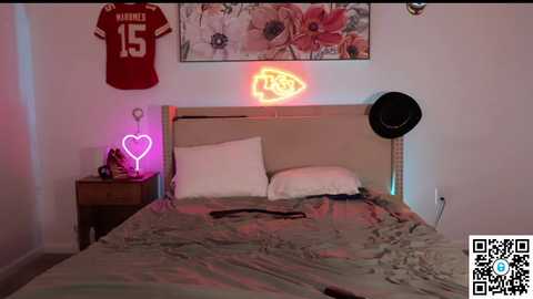 A video of a cozy bedroom with a bed, a neon heart, a red jersey, floral artwork, and a black hat, all bathed in soft pink and purple lighting.