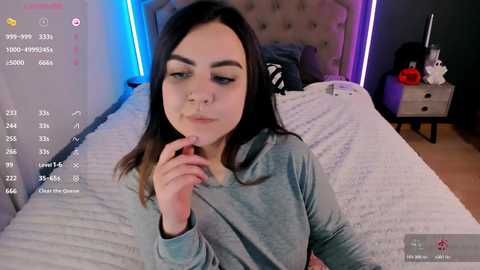 Video of a young woman with dark hair, light skin, and a septum piercing, sitting on a bed with a white quilt, in a dimly lit room with neon lights.