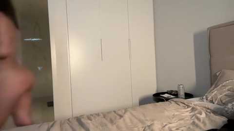 A video of a dimly lit bedroom with a white wardrobe, beige bed, and a person's arm blurred in the foreground.