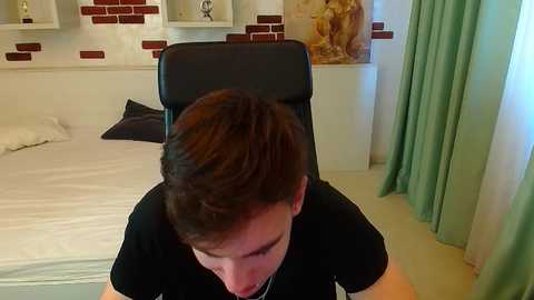 A video of a young man with short brown hair, wearing a black t-shirt, sitting in a black office chair in a modern bedroom with white walls, green curtains, and a brick-patterned accent wall.