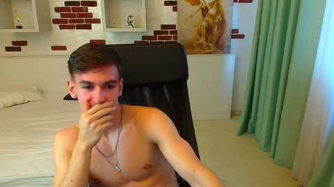 Video of a shirtless, slim, young man with short brown hair, covering his mouth with his hand, sitting on a black chair in a modern, brick-wall decorated bedroom with white and teal accents.