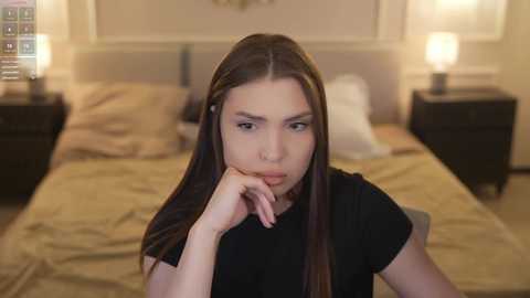 Video of a young woman with long brown hair, wearing a black shirt, resting her chin on her hand, looking contemplative in a softly lit bedroom with beige bedding, two nightstands, and a wall-mounted light fixture.