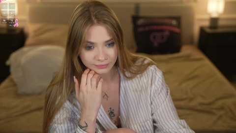 Video of a young, light-skinned woman with long, straight blonde hair, wearing a white pinstriped shirt, resting her chin on her hand in a softly lit bedroom with beige bedding.