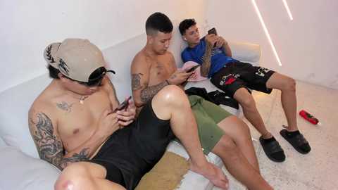 Video of three shirtless men lounging on a white couch in a minimalistic room, watching a smartphone; one has tattoos, another wears a cap, and the third in a blue shirt.