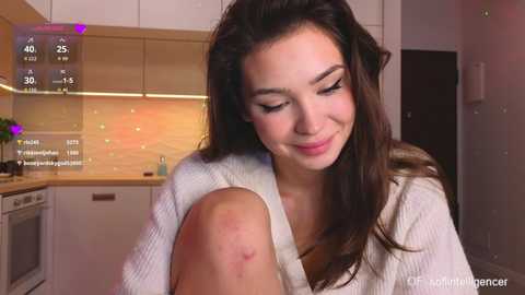 Video of a young woman with long, dark hair, wearing a white knitted sweater, sitting on a kitchen counter, with a visible bruise on her leg. Background includes a modern kitchen with white cabinets and a light-colored backsplash.