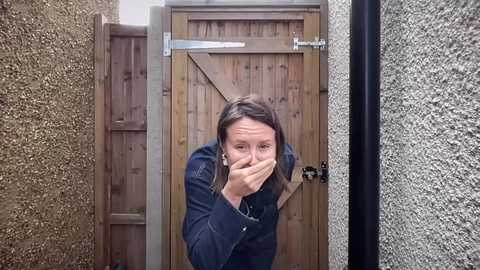 A video shows a woman with long brown hair, wearing a denim jacket, hiding her face with both hands as she stands in a narrow, textured hallway, leading to a wooden door with a metal latch.