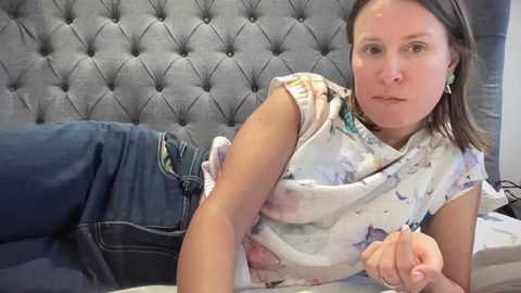Media: Video of a middle-aged woman with short brown hair, wearing a floral blouse and jeans, lying on a bed with a tufted grey headboard, looking contemplative.