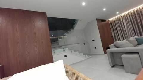 Video of a modern living room with a light gray sectional sofa, wooden paneling, glass railing staircase, and beige curtains.