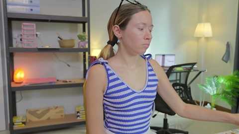 Media: Video of a fair-skinned woman with blonde pigtails in a blue and white striped tank top, sitting in a modern office with shelves, plants, and a lit lamp.