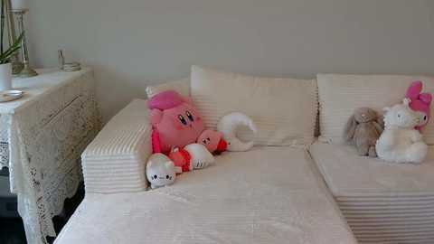 A video of a beige, striped couch with two plush toys resembling Kirby and a white bear, placed against a plain, light gray wall.