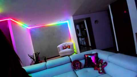 Video of a modern, dimly lit living room with a white sectional sofa adorned with colorful pillows. Neon rainbow lighting accents the ceiling, contrasting with a minimalist, white-walled setting.