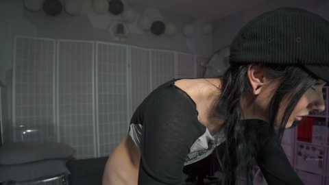 Video of a woman with long black hair, wearing a black hat and off-shoulder black top, leaning forward in a dimly lit room with white blinds and a metal fan.