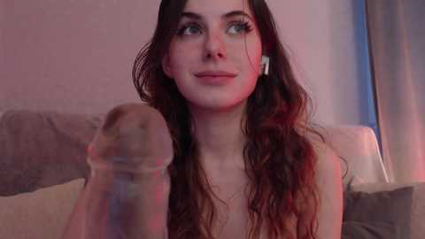 Video of a young, fair-skinned woman with long, wavy brown hair, wearing headphones, sitting on a bed, looking up with a neutral expression. The background features a beige wall and a grey blanket.