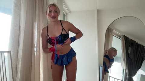Video of a smiling, blonde woman in blue lingerie with a short skirt and matching accessories, standing in front of a mirror in a softly lit room.