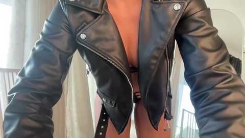 Video of a person wearing a black leather jacket, open to reveal a low-cut, black lace bra. The background shows a modern, minimalist room with light-colored walls and a window.