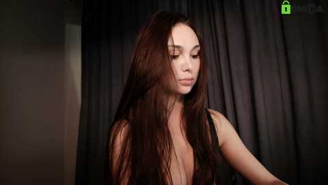 Video of a young woman with long, straight brown hair, fair skin, and slender physique, wearing a black dress, standing against dark, draped curtains. She has a serene expression.