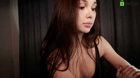 Video of a young Asian woman with long, straight black hair, wearing a black top that reveals her cleavage, against a dark curtain background.