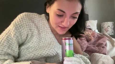 Video of a young woman with long dark hair, wearing a textured, light-colored sweater, reading a magazine while sipping from a green and white can labeled \"Fanta.\" The background features a bed with floral pillows and a black lampshade.