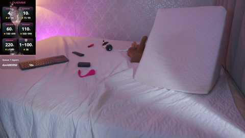 A video of a plush toy resembling a cat with a white body and pink nose, lying on a white bed, surrounded by a dimly lit, soft-focus room.